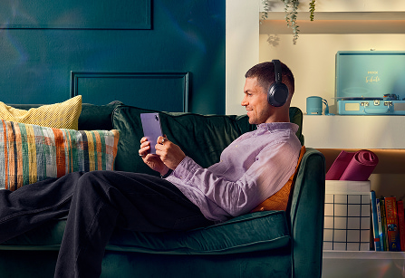 A man sitting comfortably on a green velvet sofa, wearing over-ear headphones and looking at a purple tablet.