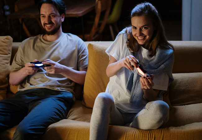 A smiling young couple sitting on a couch together, playing video games with wireless controllers.