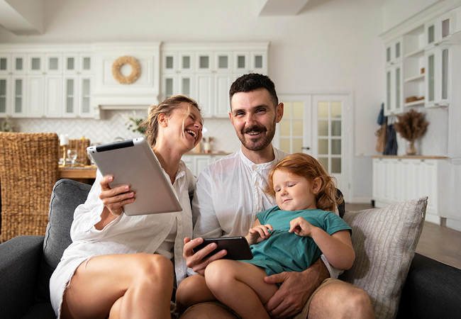 A family of three sitting on a couch in a modern, bright kitchen-living area. A woman laughs while holding up a tablet, a man smiles at the camera holding a smartphone, and a young child in a green dress sits on his lap watching the smartphone screen.