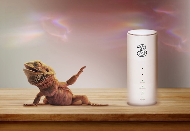 A bearded dragon rests on a wooden wooden surface next to a white Three 5G/broadband router. The lizard has its eyes closed, mouth open, and one leg raised in a dramatic, blissful pose. Warm, rainbow light flares float in the soft background.