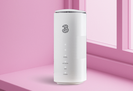 A front of a white Three 5G Broadband Hub (router) standing on a pink windowsill with a pink window frame, matching the wall colour. The '3' logo is at the top of the hub and there is a column of indicator lights, from top to bottom: Signal, Network, Wifi, and Power.