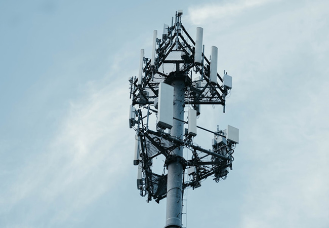 A low-angle shot of a telecommunications tower against a pale blue, wispy sky. The top of the metal pole is densely packed with various white cellular antennas and rectangular panels mounted on a triangular framework, representing modern mobile network infrastructure.