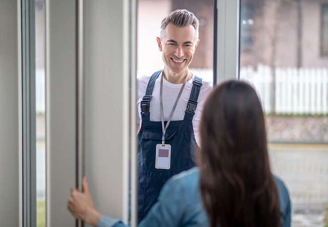 A smiling technician in dark blue overalls stands at a glass door, wearing an identification badge around his neck. He is being greeted by a woman (viewed from behind) as he arrives for a home installation appointment.