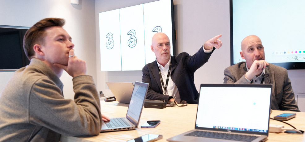 Three people seated at a table, having a meeting, each using a laptop, engaged in discussion or collaboration.
