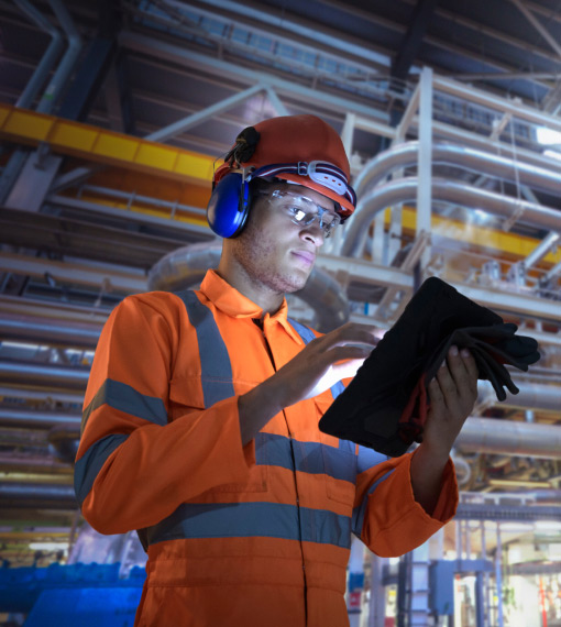 Man in hard hat, safety glasses, and hearing protection using a tablet in an industrial setting with pipes.