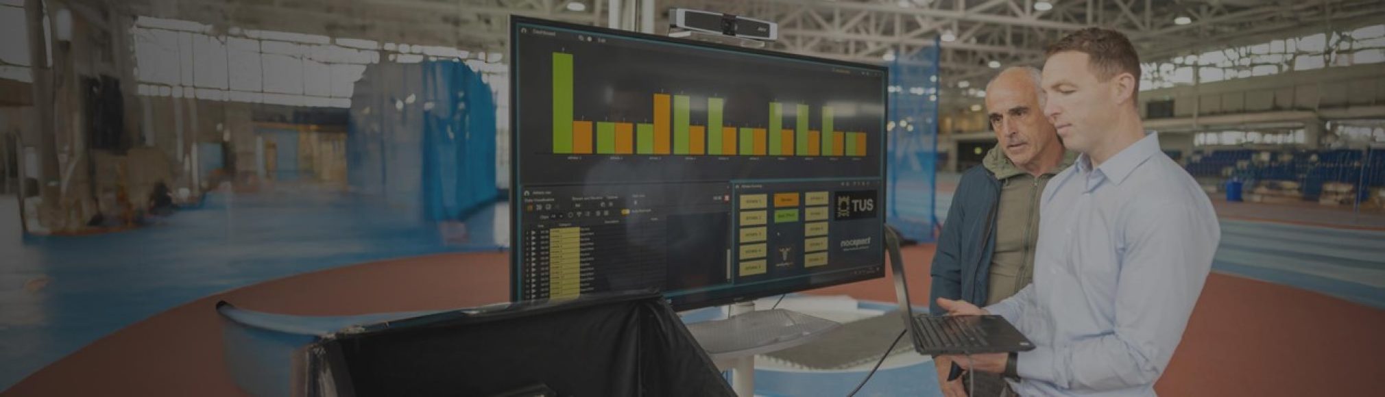 Two men stand in a large industrial space, looking at a large monitor displaying graphs and data. The man on the right, holds a laptop and is actively engaged with its screen. The man on the left is looking at the large monitor.
