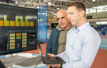 Two men stand in a large industrial space, looking at a large monitor displaying graphs and data. The man on the right, holds a laptop and is actively engaged with its screen. The man on the left is looking at the large monitor.