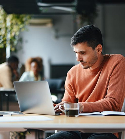 A person is seated at a desk in an office, focused on a laptop, on the desk is also a cup of coffee and some paperwork.