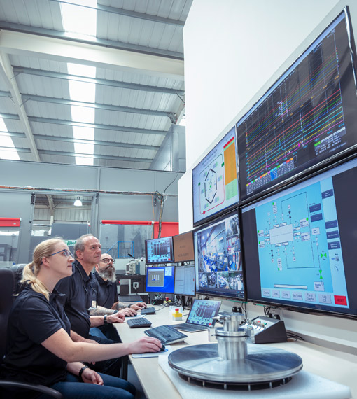 Three people in a large workplace sitting at a desk, looking at a number of large screens