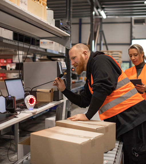 Two workers in a warehouse in front of a conveyor belt with boxes, they are wearing hi-vis vests. One worker is using a PA microphone