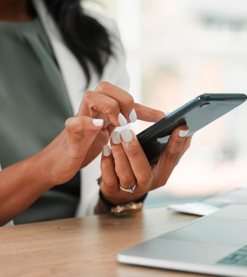 A person sitting at a desk, focused on her phone, with a laptop beside them.