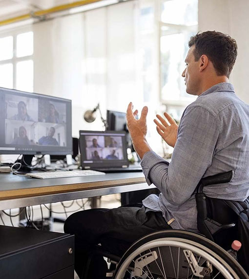 A person sits at a desk, in an online meeting on two computer monitors in a well-lit office environment.