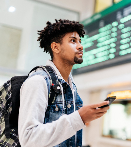 A young man with a backpack looks up at a departure board while holding a smartphone.