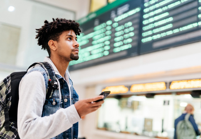 A young man with a backpack looks up at a departure board while holding a smartphone.