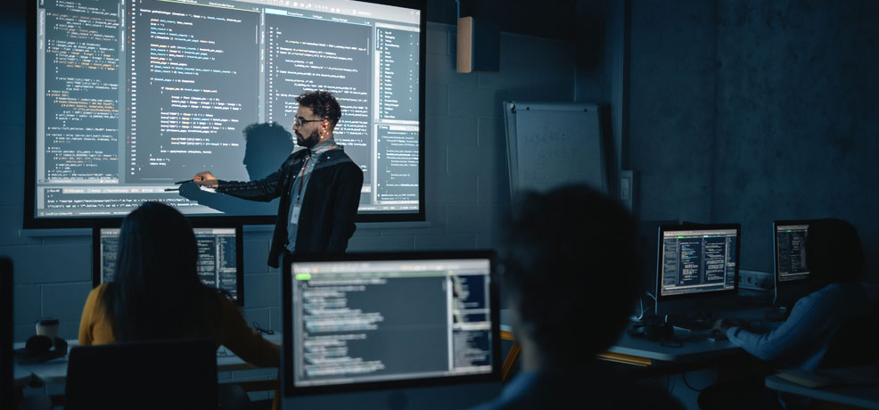 Teacher stands in front of a large screen displaying data in a classroom setting surrounded by students sitting in front of computers.