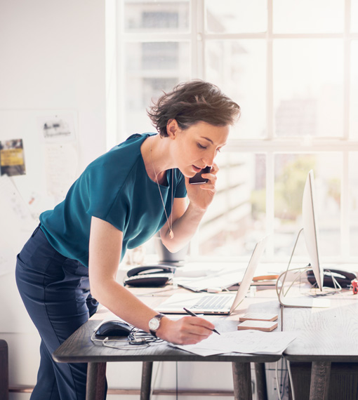 Person stands at a desk with a laptop open with phone in hand, taking notes
