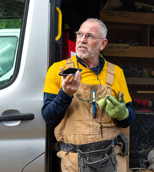Person standing in front of an open work van, wearing overalls and a toolbelt talking on the phone.