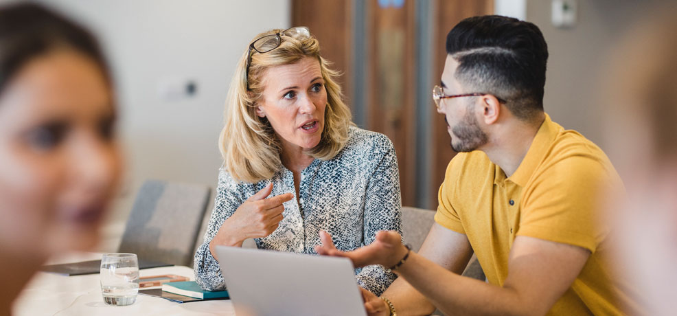 Two people sitting at a conferance table in front of a laptop, engaged in conversation.