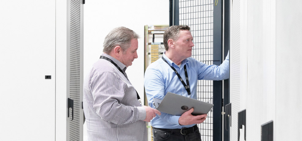 Two people are in a room full of servers, examining a computer together, engaged in conversation.