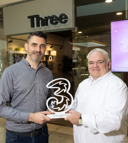 A shot of a man presenting a white Three award to another man, standing in front of a Three store.