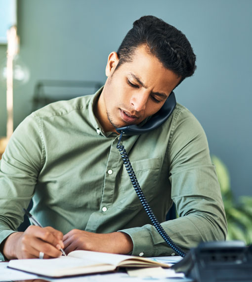 Person sitting at a desk, talking on a landline phone taking notes.