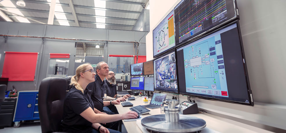 Three people in a large workplace sitting at a desk, looking at a number of large screens