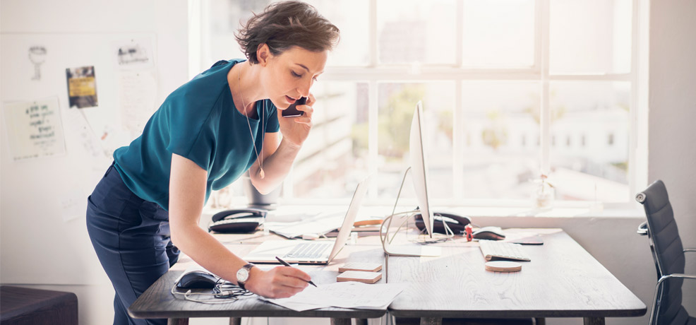 Person stands at a desk with a laptop open with phone in hand, taking notes
