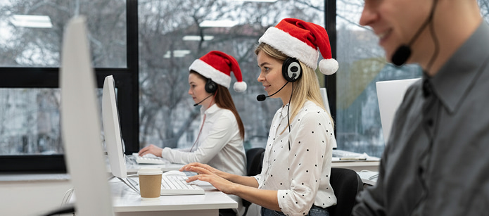Customer service representatives in a call center, wearing Santa hats and headsets, working at computers in front of a window with a snowy view.
