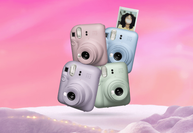 Four Fujifilm Instax instant cameras stacked together in pastel colors: pink, blue, purple, and green. The blue camera is ejecting a photo of a person blowing a bubble.