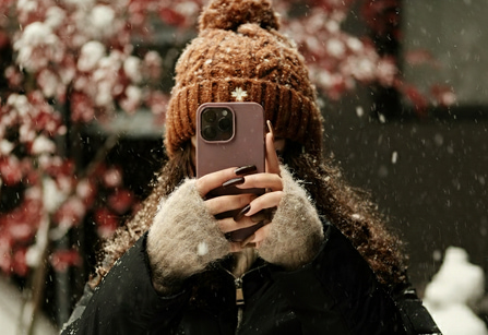 Person with curly hair and a brown knit hat taking a photo with a smartphone in the snow.