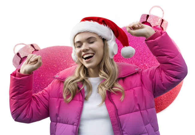 Smiling young woman wearing a Santa hat, a pink puffer jacket, and white over-ear headphones, dancing against a backdrop of two large, sparkly pink Christmas ornaments.