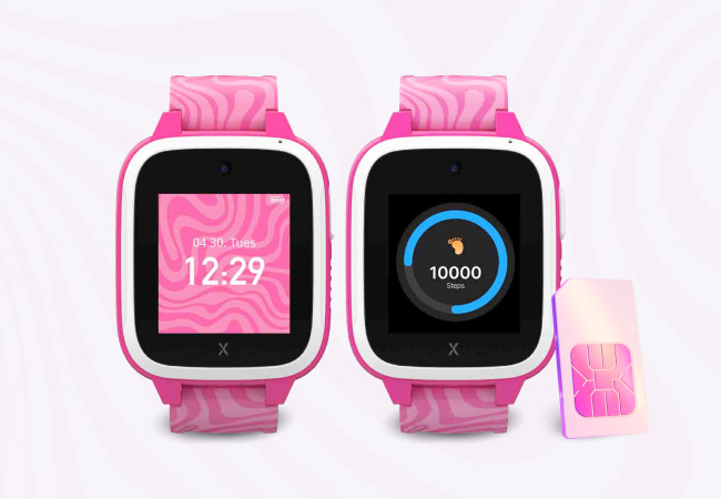 Two vibrant pink Xplora children’s smartwatches are displayed side-by-side against a pale pink wavy background. The watch on the left shows a digital clock at 12:29, while the one on the right displays a 10,000-step goal. A matching Three pink SIM card rests to the right of the devices.