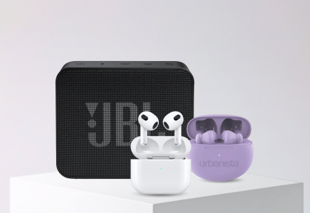 A collection of audio devices on a white block against a plain grey background. A black JBL speaker. In front of the speaker are a pair of white Apple AirPods in their open charging case, and a pair of purple Urbanista earbuds in their open case.
