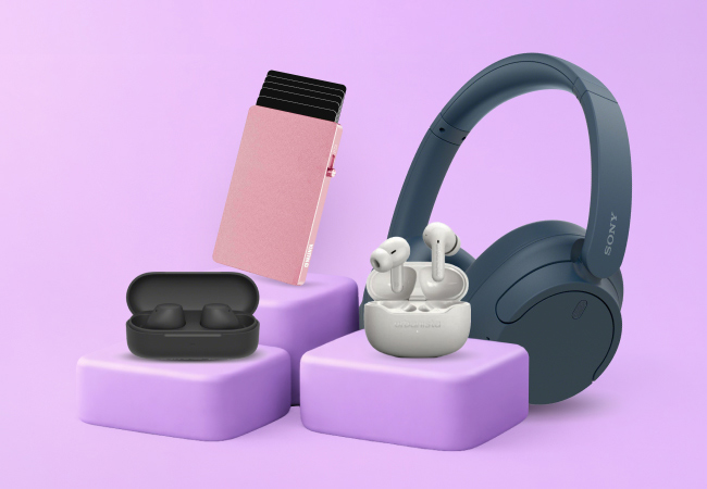 A collection of tech accessories including navy headphones, white earbuds, a black earbud case, and a pink card protector, all displayed on purple tiered platforms.