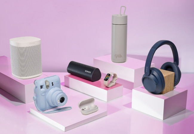 A collection of modern tech gadgets and a water bottle arranged on pink and white geometric pedestals against a lilac background.