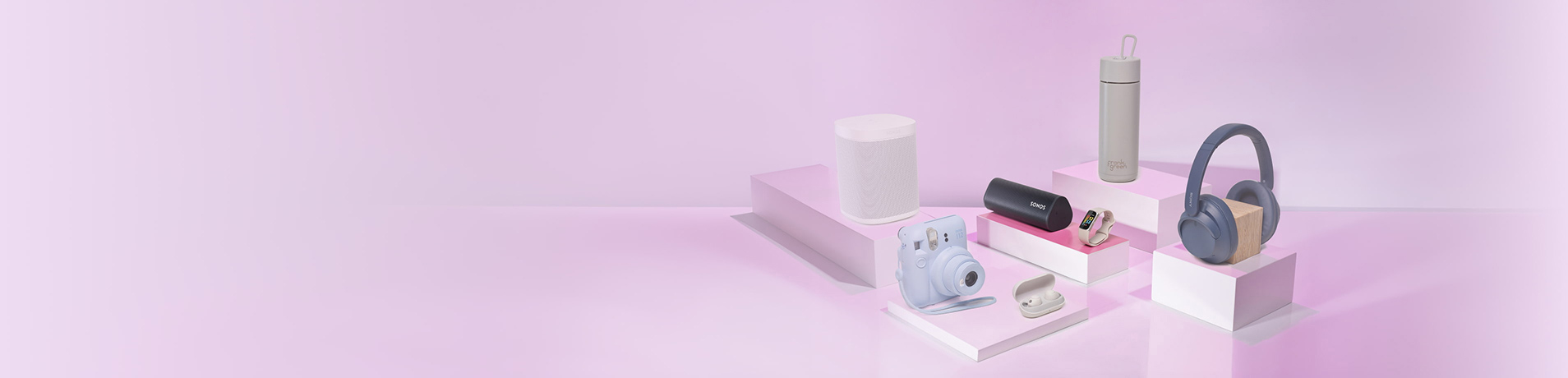 A collection of modern tech gadgets and a water bottle arranged on pink and white geometric pedestals against a lilac background.