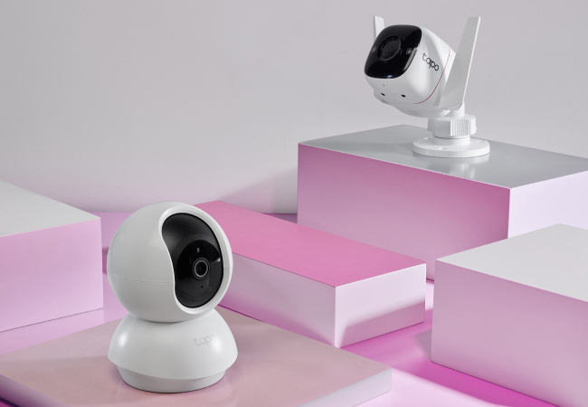 Two white TP-Link Tapo security cameras, one indoor pan-tilt model and one outdoor bullet model with antennas, displayed on pink and white geometric blocks.