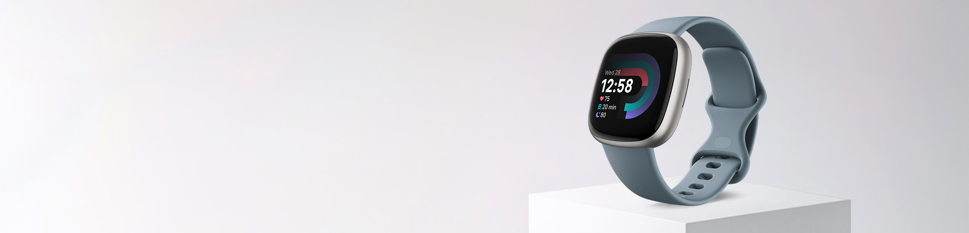 A close-up of a Fitbit Versa 4 smartwatch with a sleek, silver-toned frame and a blue-grey band, displayed on a white rectangular pedestal against a plain white background. The watch face shows "12:58" and colorful health tracking graphics.