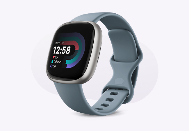 A close-up of a Fitbit Versa 4 smartwatch with a silver-toned body and a blue-grey silicone band, displayed on a light, subtly textured background. The watch shows the date and time along with colorful icons and progress bars showing health metrics