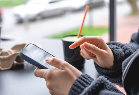 Close-up shot of a person wearing a dark, textured fleece jacket using a smartphone. One hand holds the phone while the index finger of the other hand points at the screen. In the blurred background, a black takeaway cup filled with a green drink and an orange straw sits on the table, overlooking a street scene.