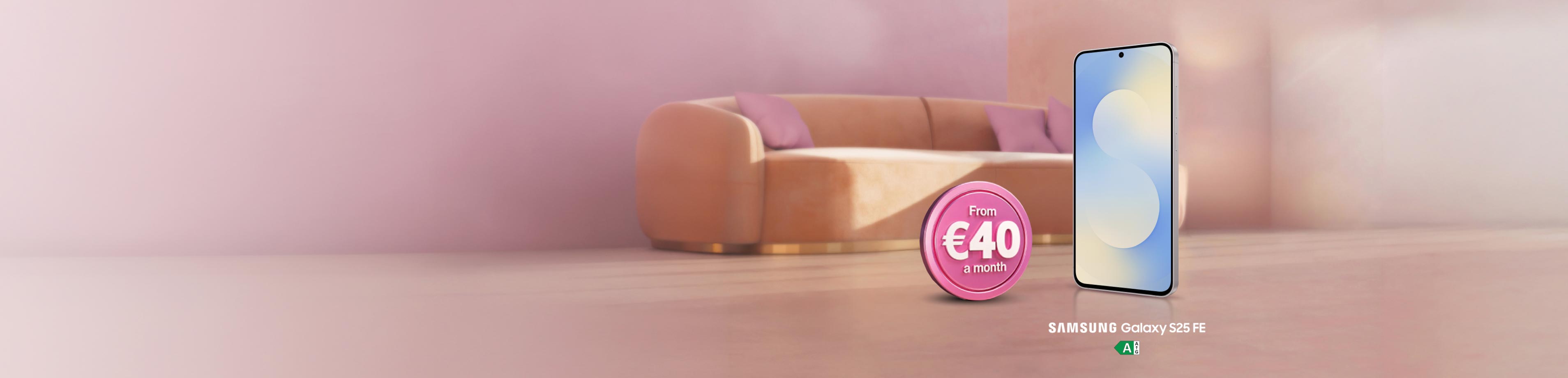 Promotional image for the Samsung Galaxy S25 FE smartphone, standing upright against a warm, pink-toned indoor background with a blurry couch. Next to the phone is a pink token that reads "From €40 a month".