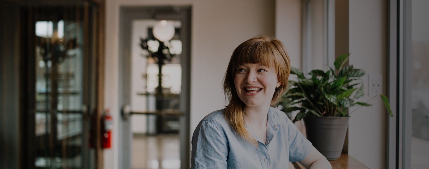A smiling person with reddish-blonde hair and bangs looks away from the camera, sitting by a window with a plant.