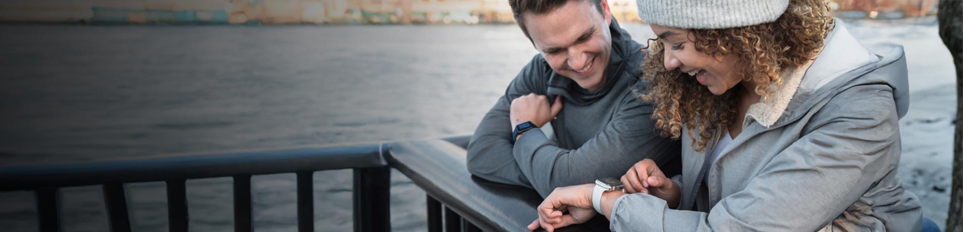 Two people are leaning against a black railing beside a body of water. They are looking down at a white smartwatch on one of their wrists.