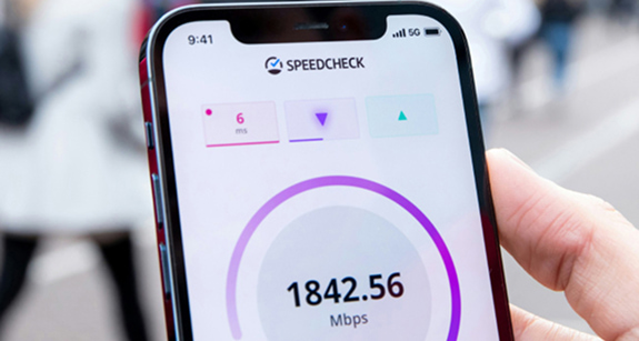 Close-up shot shows a hand holding a smartphone. A speed test app shows a download speed of 1842.56 Mbps and a latency of 6 ms. The phone's status bar indicates a 5G connection. A blurred, urban background is visible behind the hand.