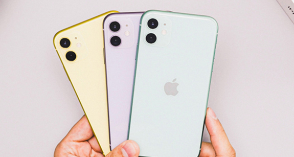 A person's hands are holding three iPhones in a fan-like arrangement against a light gray background, they are in different pastel colors: yellow, purple, and mint green.