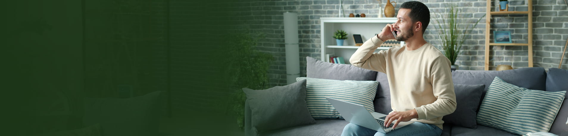 A man sits on a grey sofa in a modern living room, talking on a smartphone and using a laptop. The image features a dark green color gradient on the left side, transitioning into the brightly lit scene of the man working.