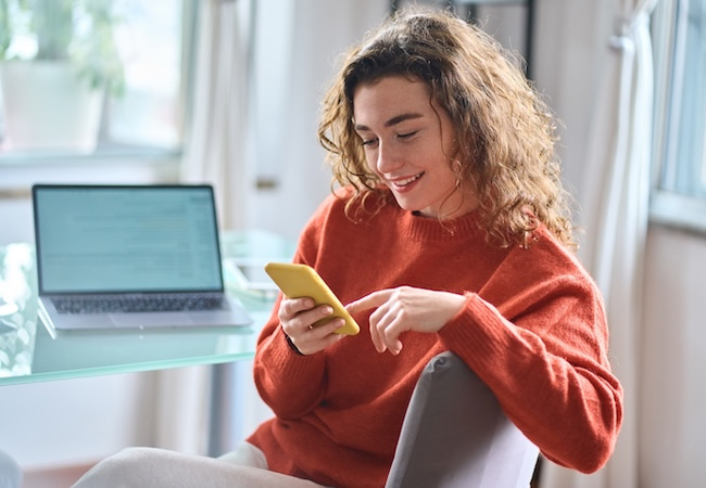 A woman with curly hair sits at a glass table, smiling as she looks at her yellow smartphone. A laptop is open on the table behind her in a brightly lit, modern room.