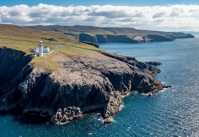 An aerial view of a white lighthouse standing on a steep, grassy cliff edge overlooking a vast blue ocean. Rugged, rocky coastline and rolling hills extend into the distance under a bright, partially cloudy sky.