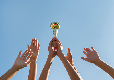 Hands holding up a trophy against a blue sky in celebration of a team victory.