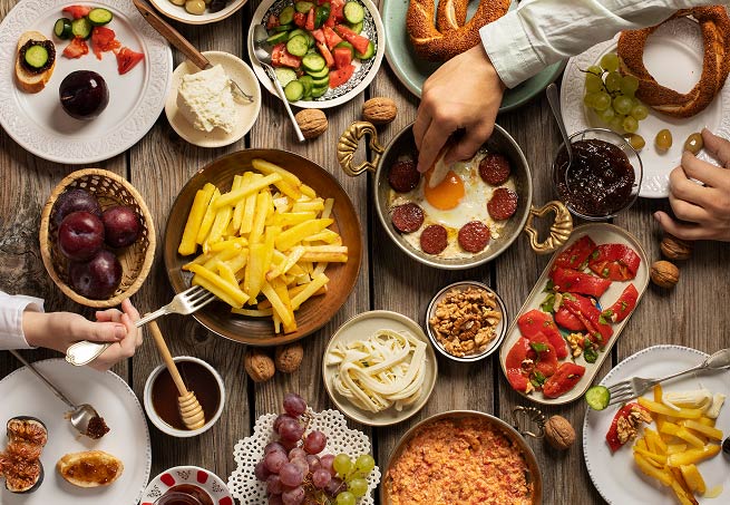 Overhead view of a large spread of breakfast dishes on a wooden table, with hands reaching for food